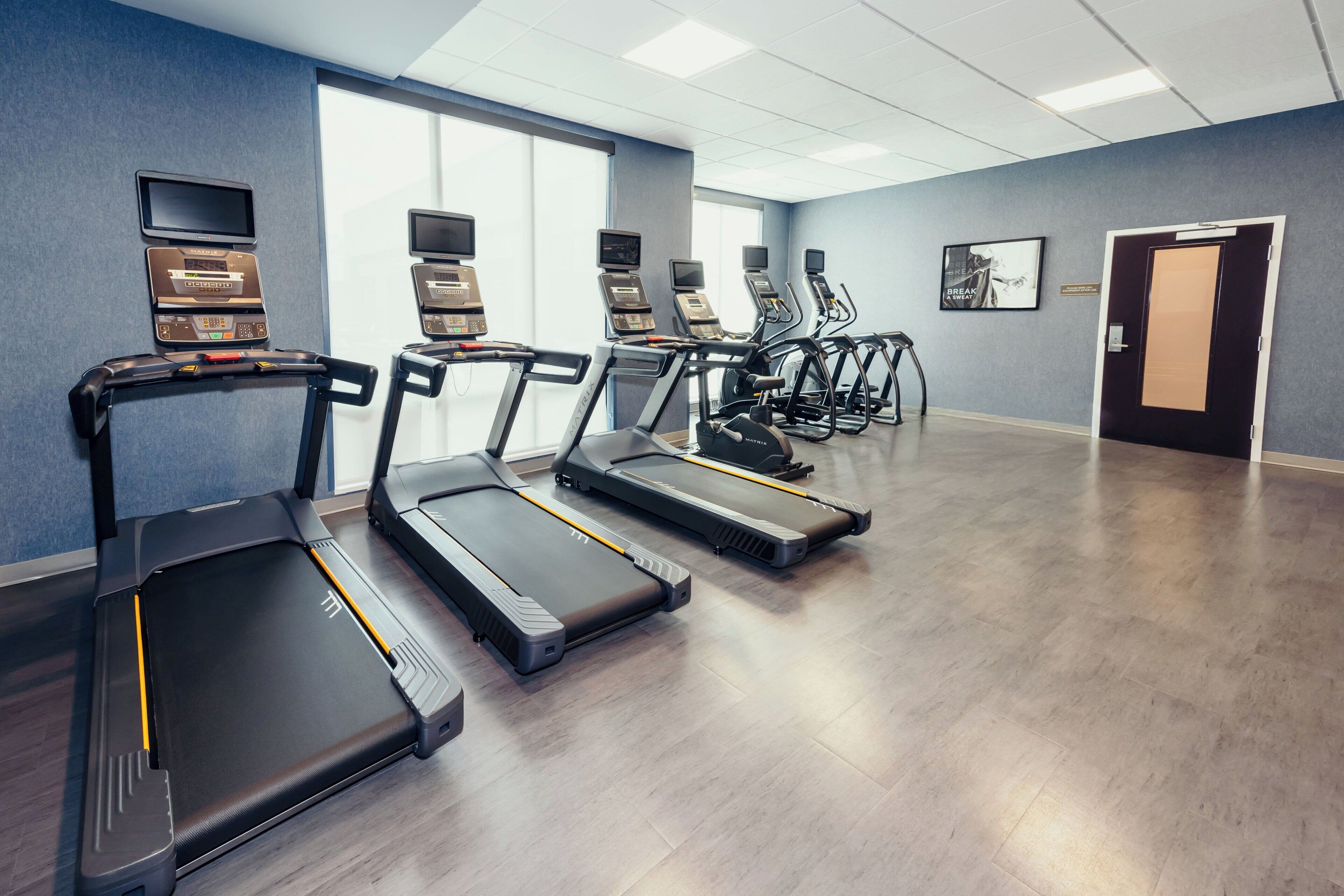 A fitness center with treadmills and elliptical machines, a large window, and a door with a sign.