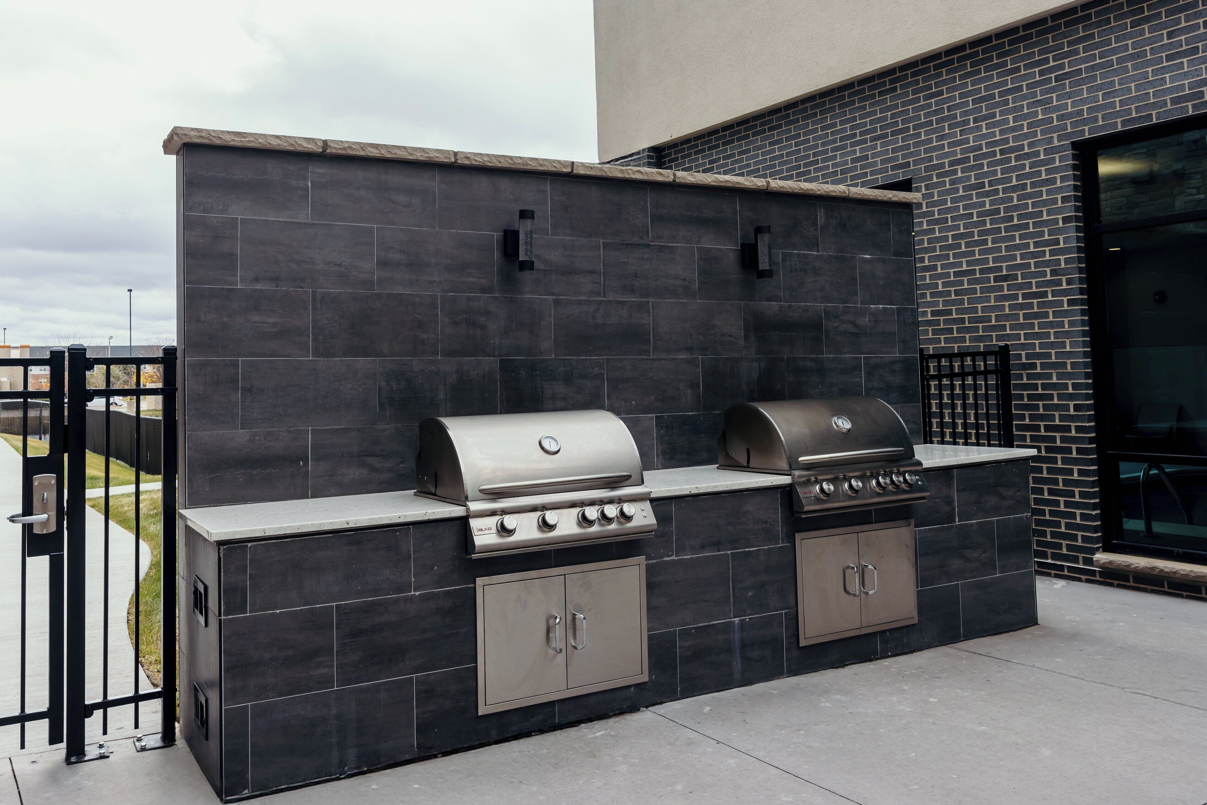 An outdoor kitchen with a grill, a built-in oven, and a sink, set against a dark tiled wall.