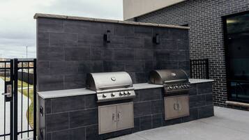Une cuisine extérieure équipée d’un barbecue, d’un four encastré et d’un évier, le tout installé devant un mur recouvert de carreaux foncés.