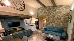 A modern living room with a sofa, a TV, a dining table, and a kitchen area visible through an open door. - Villa "terre D'azur" Avec vue mer , Piscine, Jardin Clos, Rénovée (La Croix-Valmer)