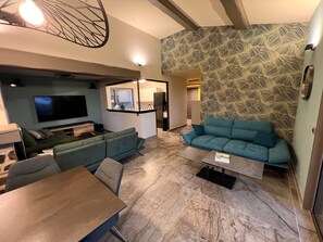 Ein modernes Wohnzimmer mit Sofa, Fernseher, Esstisch und einem durch eine offene Tür einsehbaren Küchenbereich.