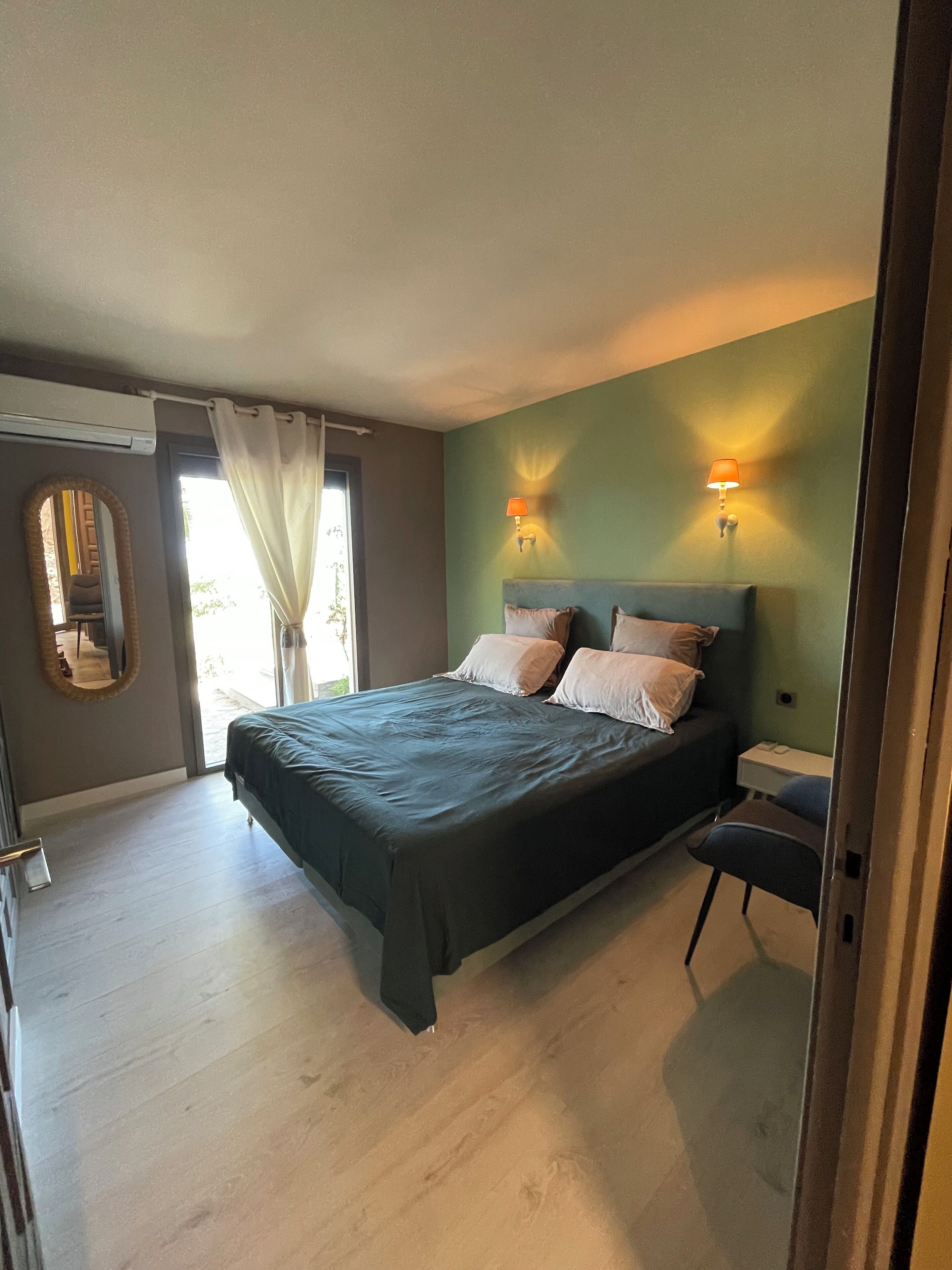 Un dormitorio con cama, mesita de noche, espejo y ventana con cortinas.