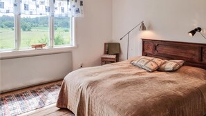 Quarto com uma janela grande, cama com almofadas estampadas, mesa de cabeceira, uma cadeira e abajur.