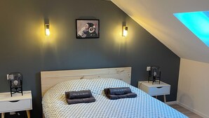 Une chambre à coucher moderne avec un plafond en pente, une couvre-lit à motifs, deux tables de chevet avec des lampes et une œuvre d’art encadrée au mur.