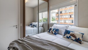 Een slaapkamer met een bed, kussens, een spiegel en een raam met uitzicht op gebouwen.