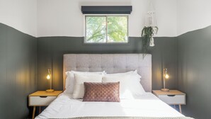 A neatly made bed with a textured pillow, two bedside tables with lamps, a window with a view of trees, and a hanging plant.