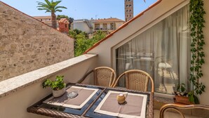 Un balcón con sillas de mimbre, una mesa y una planta en maceta. Se observa una pared de piedra y un edificio con torre al fondo.