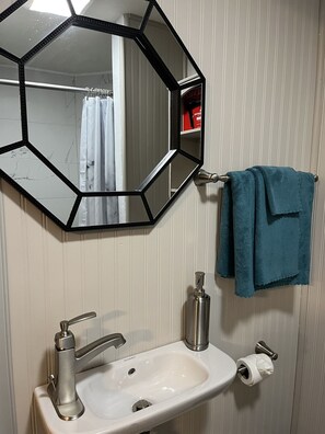 Un baño con un espejo hexagonal, un lavamanos con grifo, una toalla colgada en un perchero y un dispensador de papel higiénico.