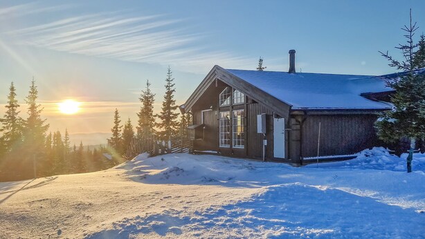 A wooden cabin with a sloped roof, surrounded by snow-covered ground and evergreen trees, with the sun setting in the background. - Beautiful home in Sjusjøen with sauna (Sjusjøen)
