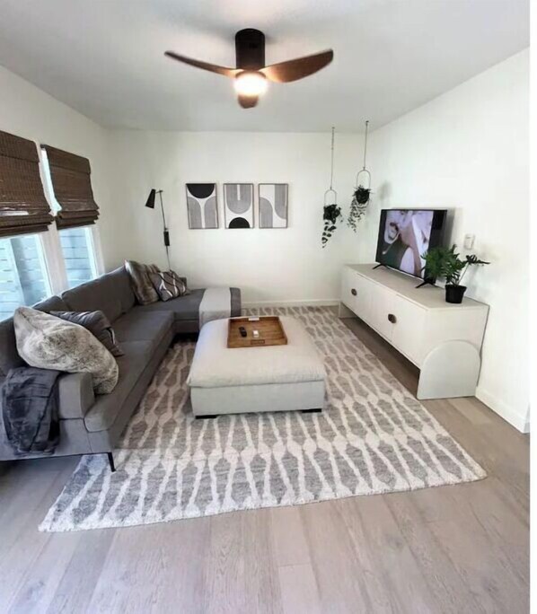 Una sala de estar moderna con un sofá gris, un otoman blanco, un ventilador de techo y un televisor sobre un mueble blanco.
