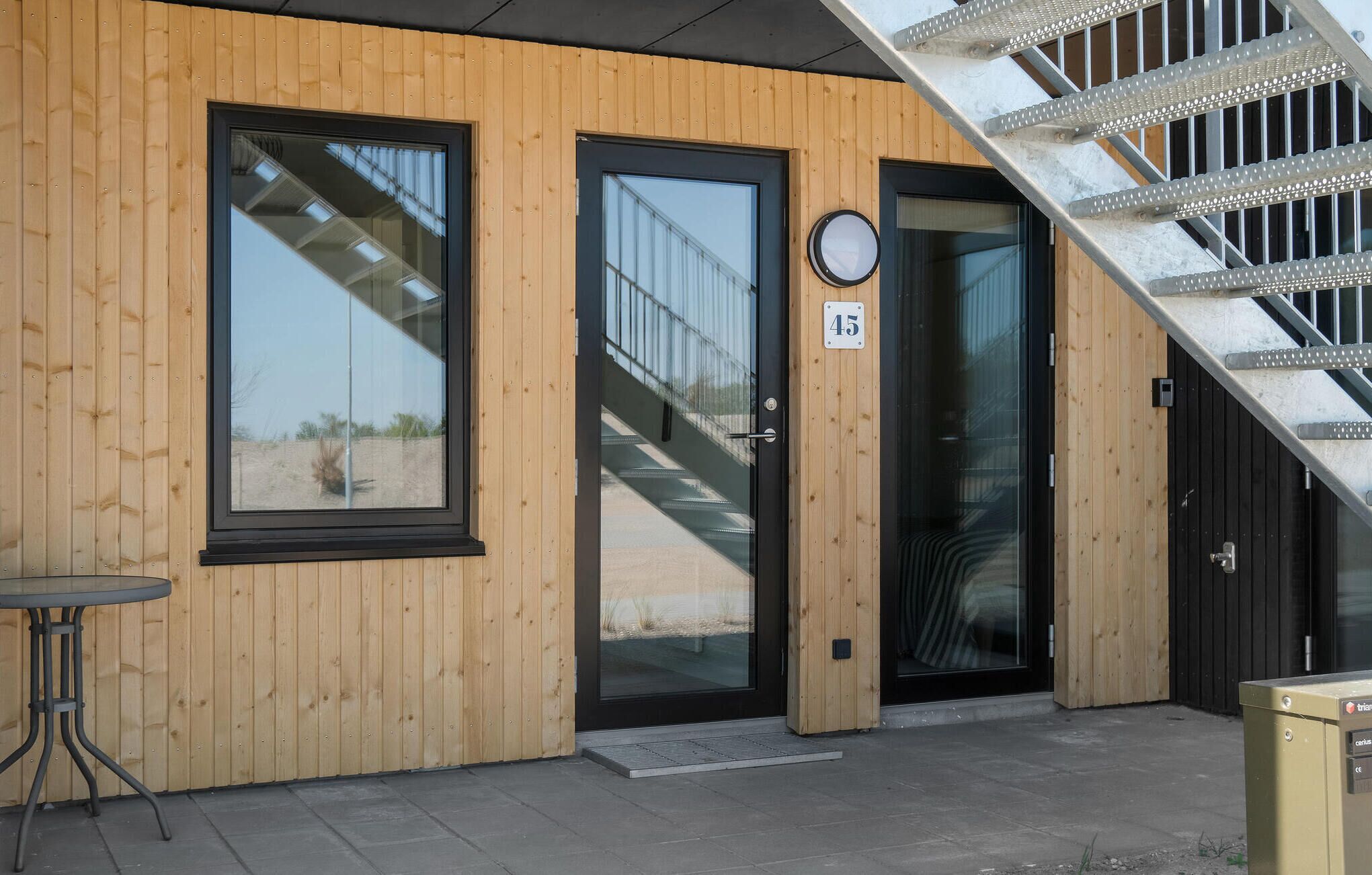 Een gebouw met houten lambrisering, twee grote ramen, een zwarte deur en een trap naar een hoger gelegen niveau.