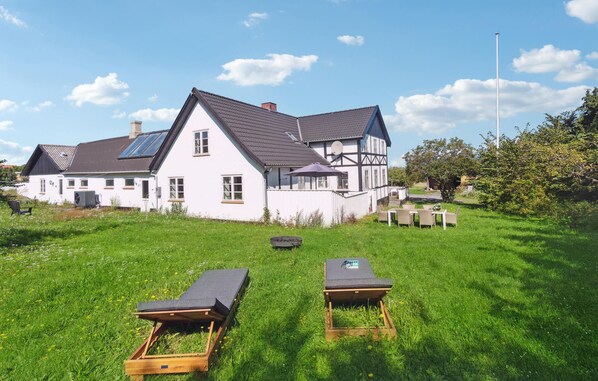 Une grande maison blanche avec un toit foncé, une antenne parabolique et une pelouse bien entretenue, agrémentée de deux chaises longues en bois.
