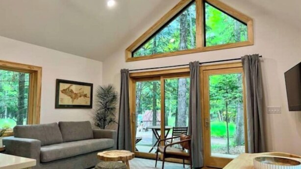 Ein modernes Wohnzimmer mit einem grauen Sofa, einem Holztisch und einem Stehocker. Große Fenster bieten einen Blick ins Freie.