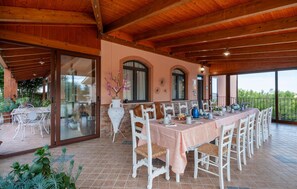 Un espace extérieur abrité, aménagé pour les repas, avec une longue table pouvant accueillir plusieurs personnes, entouré de chaises blanches à assise tressée.
