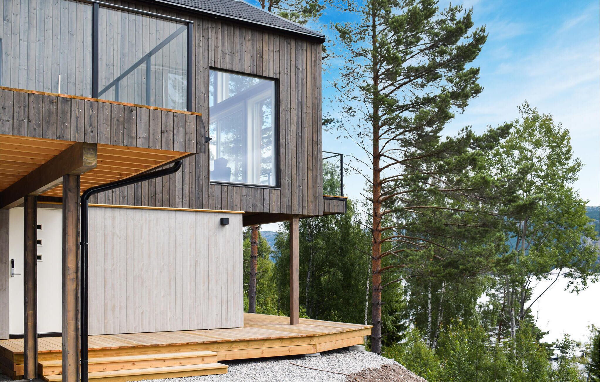 Une cabane moderne en bois, dotée d’une terrasse, de grandes fenêtres et offrant une vue sur une zone boisée.