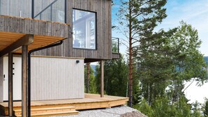 Una cabaña de madera moderna con terraza, ventanas amplias y vistas a un bosque.