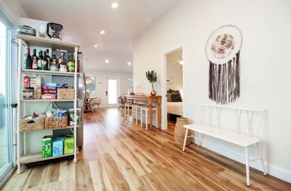 Amplia sala de estar con piso de madera, un banco blanco, una mesa de comedor con sillas, una estantería con diversos objetos y un atrapasueños en la pared.