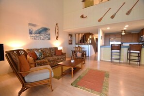 A spacious living area with a sofa, armchair, and coffee table. A kitchen is visible in the background with a microwave and chairs.
