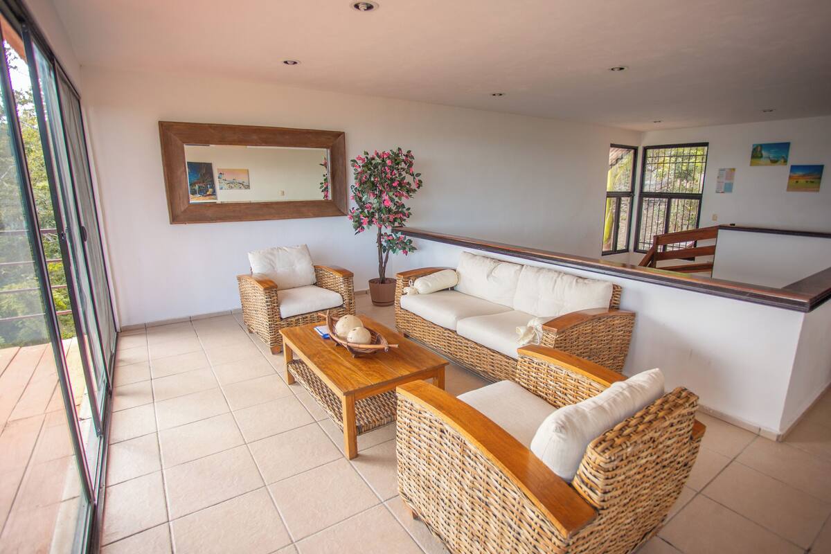 A spacious living area with a sofa, two armchairs, a coffee table, and a small side table. There is a potted plant, a large mirror, and a window with a view of greenery.