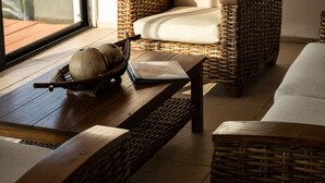 Una silla de mimbre con un cojín, una mesa de madera con un cuenco y un libro, y un ventanal con vistas a árboles.