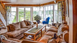 Een gezellige woonkamer met een bruine zithoek, een salontafel en een blauwe fauteuil. Grote ramen bieden uitzicht op groen buiten.