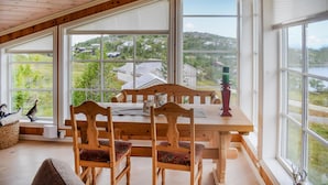 Een eetruimte met een houten tafel en stoelen, grote ramen met uitzicht op huizen en groen, en een kaars op de tafel.