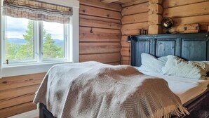 A cozy log cabin bedroom with a large window, a bed with a patterned blanket, and a dark wooden headboard.