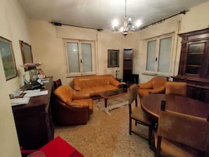 Un salon avec un canapé en cuir marron, une table basse, une table à manger avec des chaises, un lustre et deux fenêtres équipées de stores.