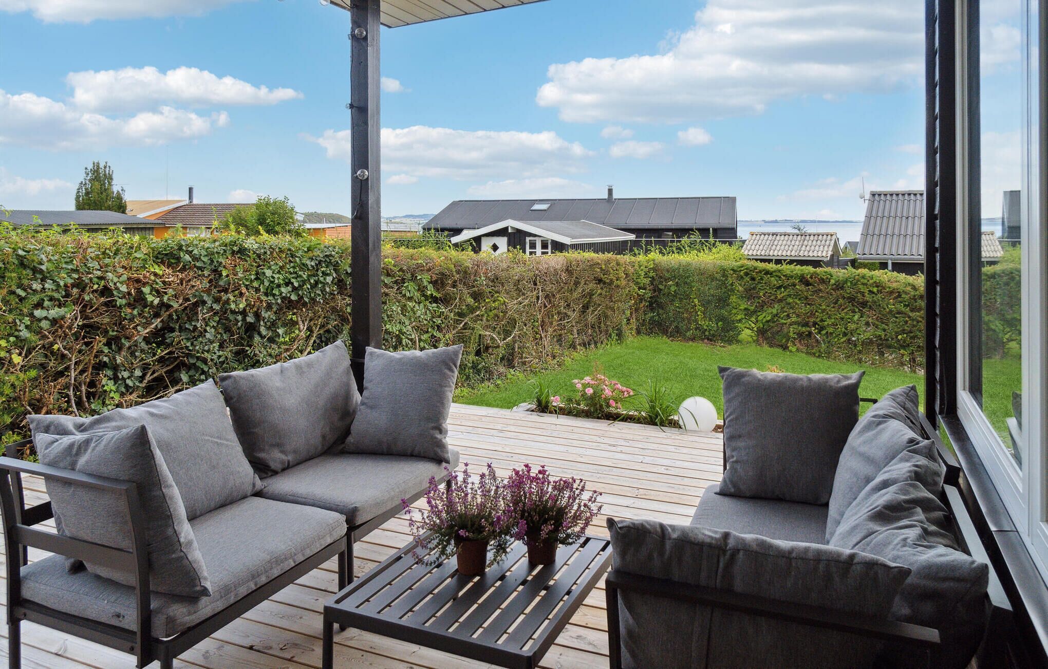 Een terras met een bank en een tafel, met uitzicht op een tuin met een haag en een huis op de achtergrond.