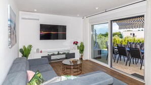 A modern living room with a grey sofa, a glass coffee table, a flat-screen TV mounted on the wall, and a sliding glass door leading to an outdoor patio with a striped awning and outdoor furniture. - Stunning home in Kastel Novi (Kastel Novi)