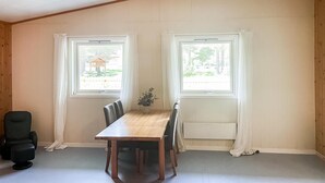 Een kamer met een houten plafond, twee ramen met gordijnen, een eettafel met stoelen, een zwarte bureaustoel en een potplant.