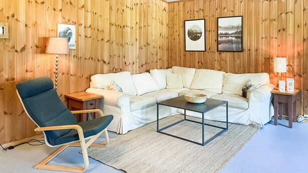 Een gezellige woonkamer met een houten lambrisering, een witte bank, een blauwe fauteuil, een salontafel en twee ingelijste foto's.