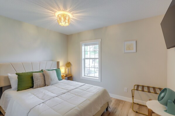 A bedroom with a large bed, a bedside table with a lamp, a window, and a wall-mounted fan.