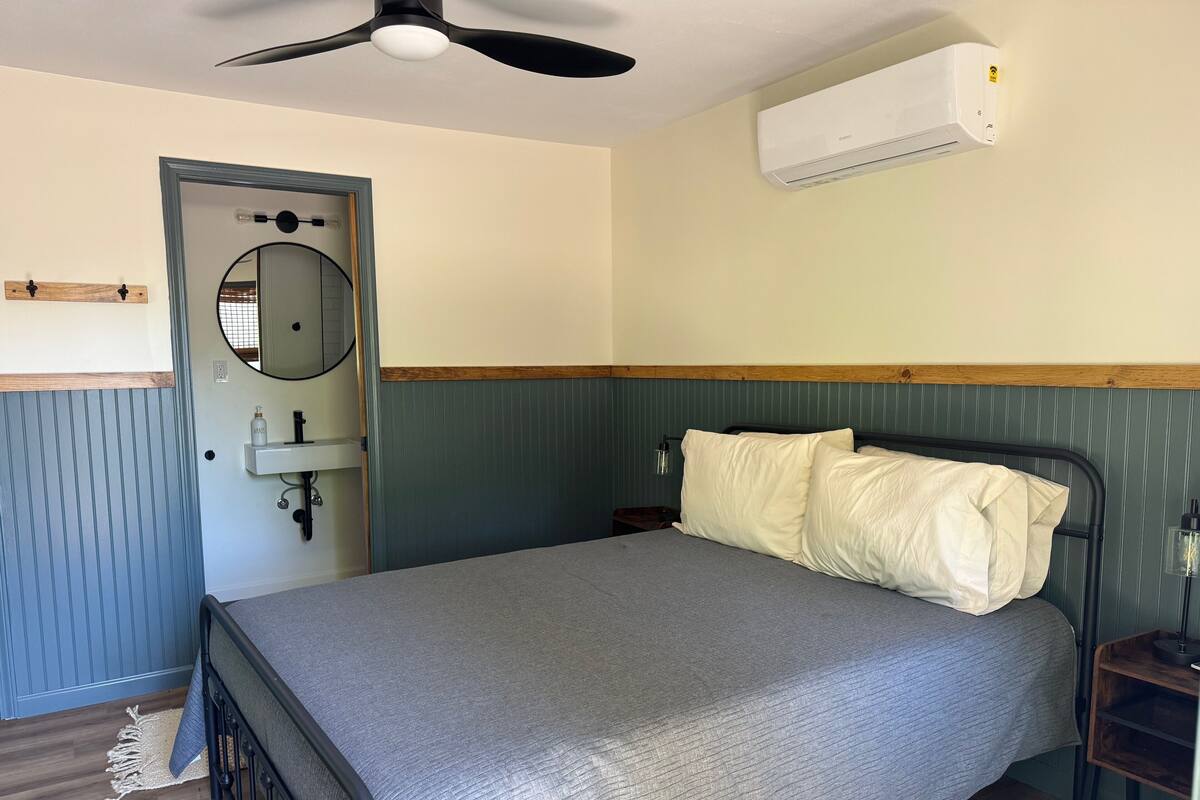 A bedroom with a bed, a ceiling fan, an air conditioning unit, a wooden door, a mirror, and a bedside table.