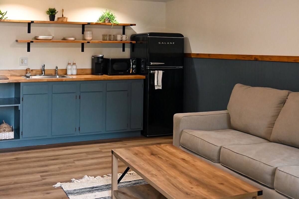 A modern living room with a wooden coffee table, a beige sofa, and a kitchen area with a black refrigerator and a microwave.