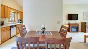A dining area with a wooden table, chairs, and a vase with flowers. There is a kitchen with wooden cabinets and a microwave. A flat-screen TV is mounted on the wall above a fireplace.