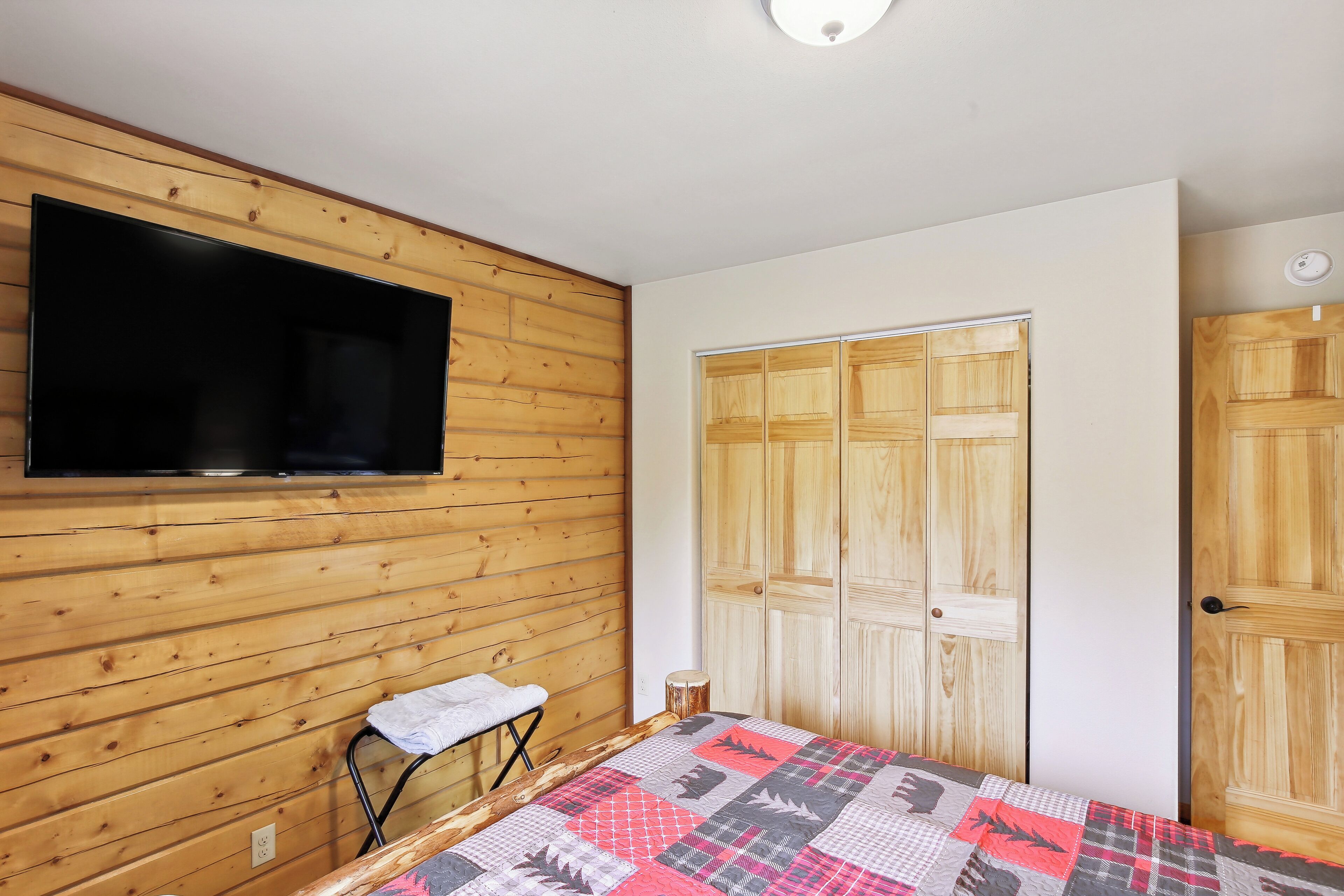 A bedroom with wooden paneling, a bed with a patterned quilt, a television mounted on the wall, and a wardrobe with a mirror.