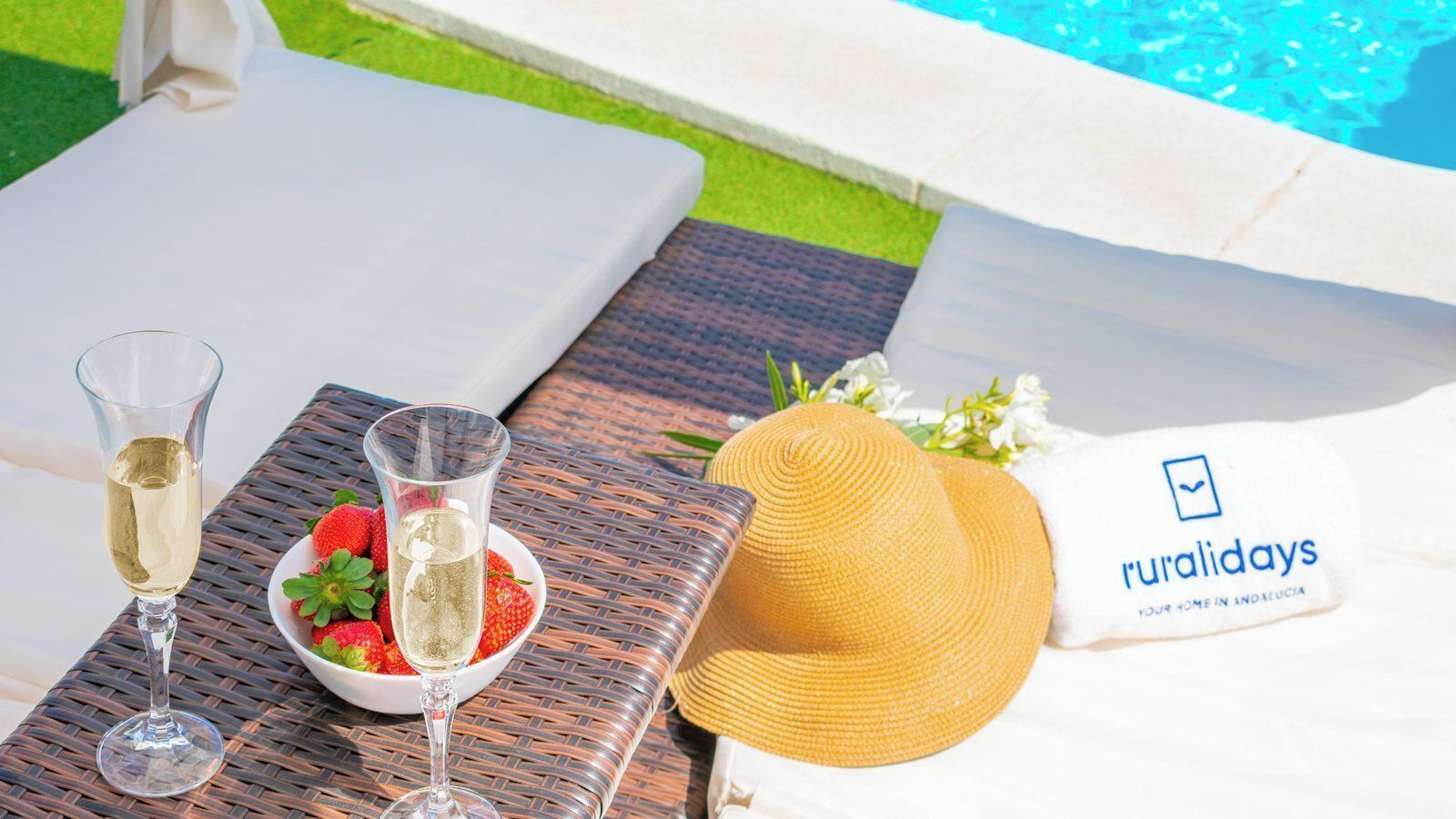 A table with a glass of white wine, a bowl of strawberries, and a straw hat with a towel.
