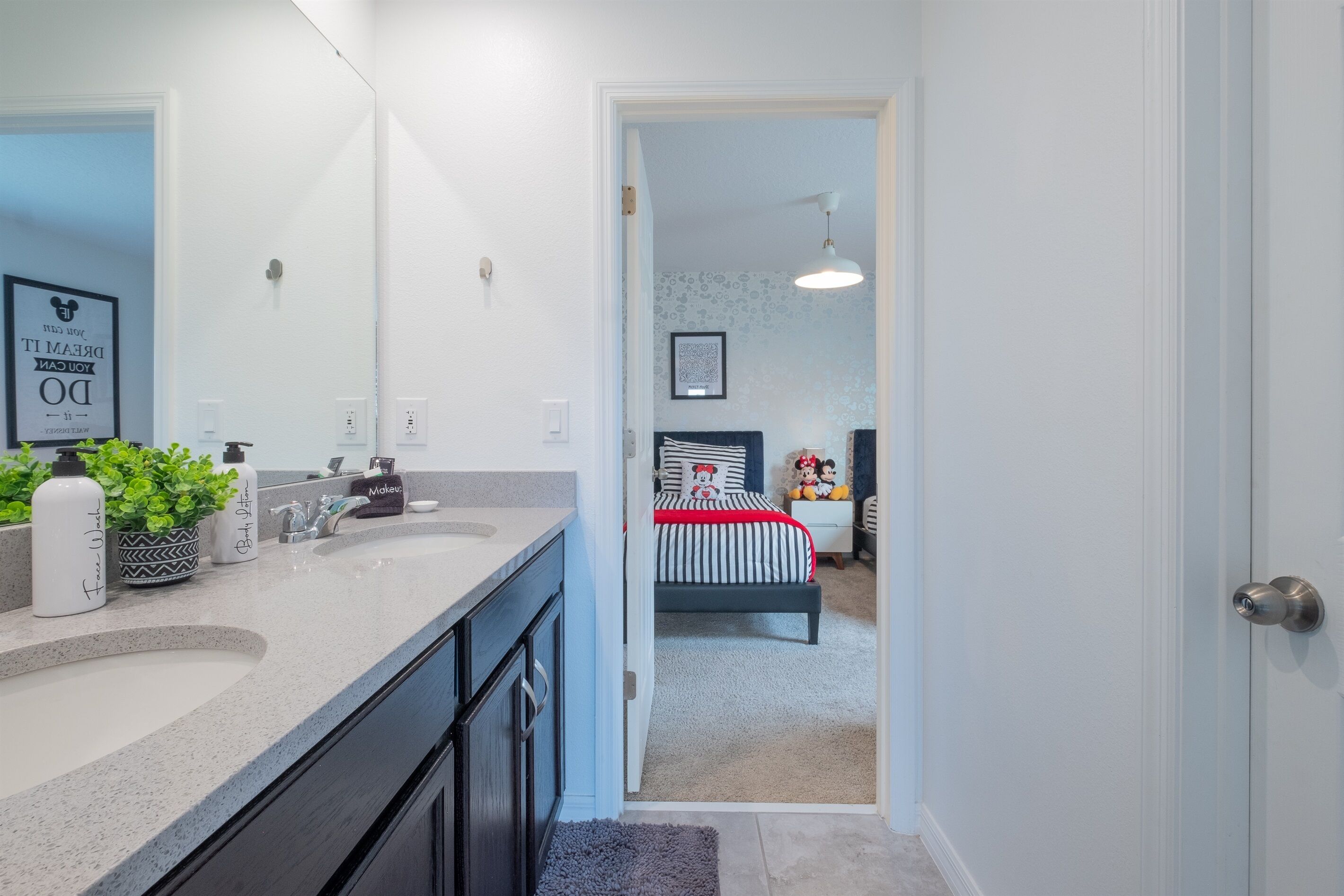 Une salle de bain moderne avec un grand miroir, un lavabo et un meuble-vasque. Une porte donne accès à une chambre à coucher équipée d’un lit et d’une table de chevet.