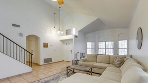 A spacious living area with a beige sectional sofa, a coffee table, and a patterned rug. A staircase with a black railing leads to an upper level. - Cookout Ready! Desert Oasis w/ Pool in Goodyear (Goodyear)