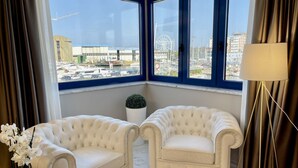 Ein Eckzimmer mit zwei weißen, gepolsterten Ohrensesseln, einer Topfpflanze und Blick auf einen Yachthafen mit Booten und einem Riesenrad.
