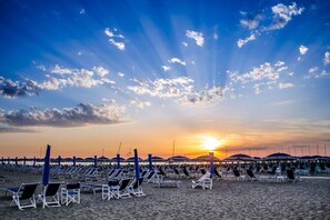A beach with rows of sun loungers and umbrellas, a pier in the background, and the sun setting over the horizon.