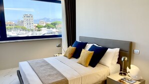 Ein modernes Hotelzimmer mit einem großen Bett, einem Nachttisch, einer Lampe und Blick auf die Stadt durch ein großes Fenster.
