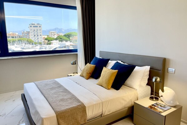 A modern hotel room with a large bed, a nightstand, a lamp, and a view of the cityscape through a large window.