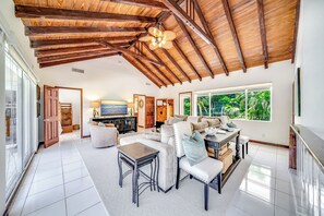 Un vaste salon doté d’un haut plafond et de poutres apparentes, d’une grande fenêtre offrant une vue sur la verdure et d’un coin salon confortable avec une cheminée.