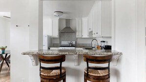 Een keuken met witte kasten, een granieten aanrecht en twee houten stoelen met geweven zittingen.