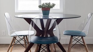 Een eetgedeelte met een ronde tafel, vier stoelen, een plafondventilator en een raam met uitzicht op bomen en een gebouw.
