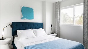 Een slaapkamer met een groot bed, wit beddengoed, een blauw hoofdeinde en een raam met patroon gordijnen.