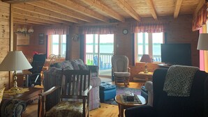 Ein gemütliches Blockhaus-Interieur mit Holzwänden, einem Essbereich, einem Wohnzimmer mit Sofa und Sesseln sowie einem großen Fenster mit Blick auf ein Gewässer.