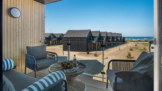 Ein Balkon mit Korbmöbeln und einem Tisch mit Früchten und einer Flasche, mit Blick auf einen Strand mit Reihenhäusern.